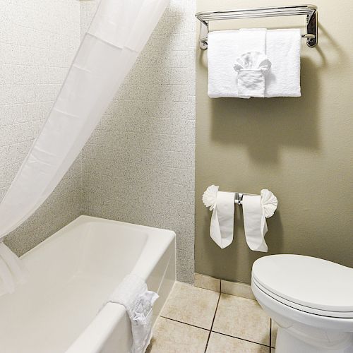 A clean bathroom with a bathtub, shower curtain, toilet, towel rack with folded towels, and a toilet paper holder on a tiled floor.