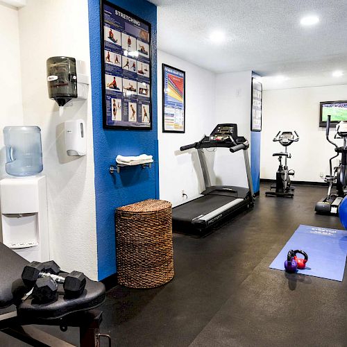 A small gym with exercise equipment, including a bench, dumbbells, treadmill, stationary bikes, yoga mat, stability balls, water dispenser, and wall mirrors.