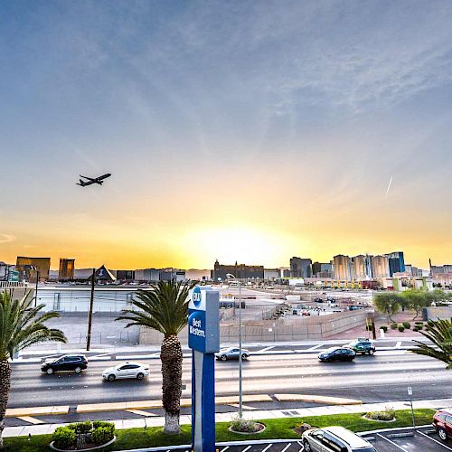 An urban landscape with a skyline at sunrise, palm trees, a road with cars, and an airplane in the sky. The skyline includes a Ferris wheel.