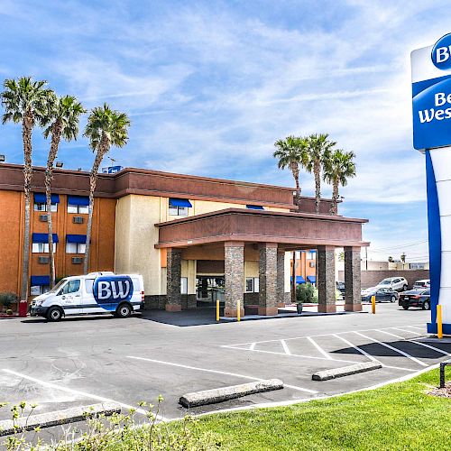 The image shows a Best Western hotel building with a parking lot in front, a BW van, palm trees, and a prominent Best Western sign beside the entrance.