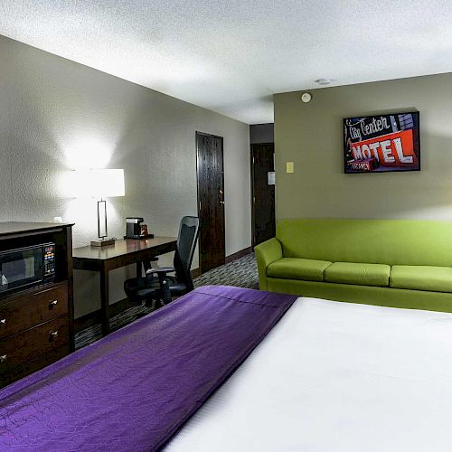 A hotel room with a bed, green couch, TV, microwave, desk with chair, lamps, and a wall sign reading 