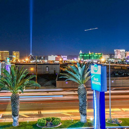 A night view of a city skyline with bright lights, palm trees, a prominent blue and white hotel sign, and a beam of light projected into the sky.