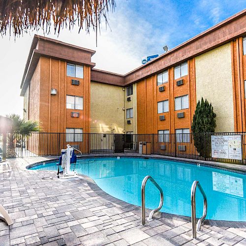 An outdoor swimming pool area with lounge chairs and a building in the background, surrounded by a fence, and clear blue skies above.