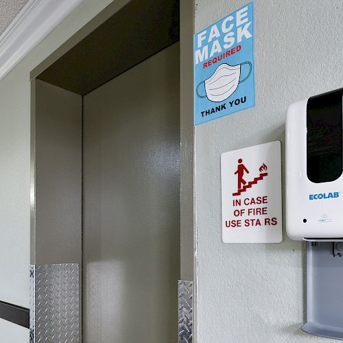 An elevator entrance with signs for mandatory face masks, fire safety, and a hand sanitizer dispenser is visible on the wall.