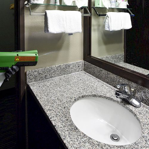 A person is seen sanitizing a bathroom sink and counter with a green handheld sprayer, next to a mirror and towels.