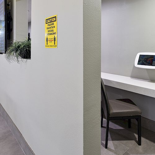 A hallway with a sign about maintaining social distancing, a small area with a desk, chair, and a tablet. The area is modern and minimalistic.