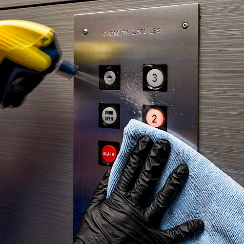 A person wearing black gloves is cleaning an elevator control panel with a microfiber cloth and a spray bottle.