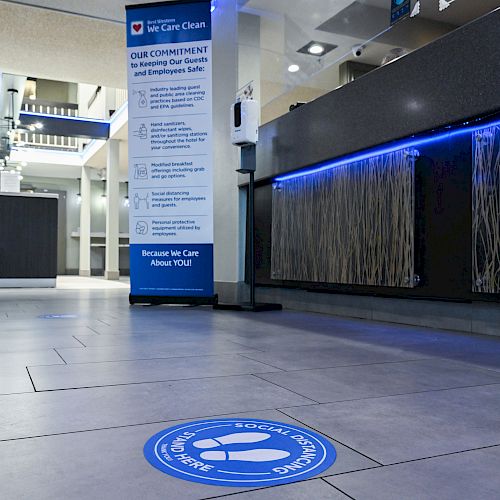 The image shows a modern lobby area with a social distancing floor marker, a hygiene commitment stand, and an illuminated reception desk.