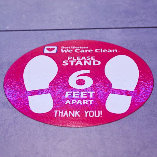 A red circle floor sticker with white footprints instructs people to stand 6 feet apart as part of health and safety guidelines. Thank you!