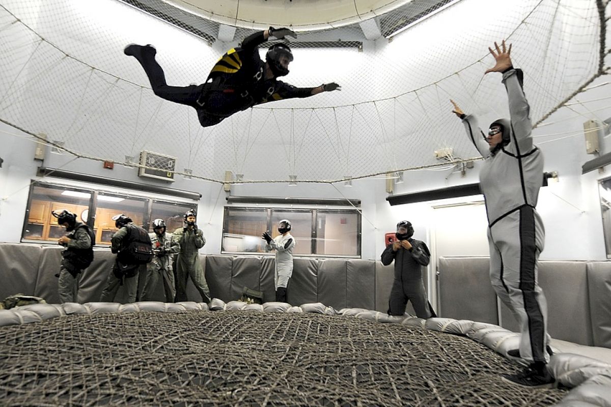 A person is floating in a wind tunnel; others in safety gear observe and take photos around the netted area, indicating an indoor skydiving facility.