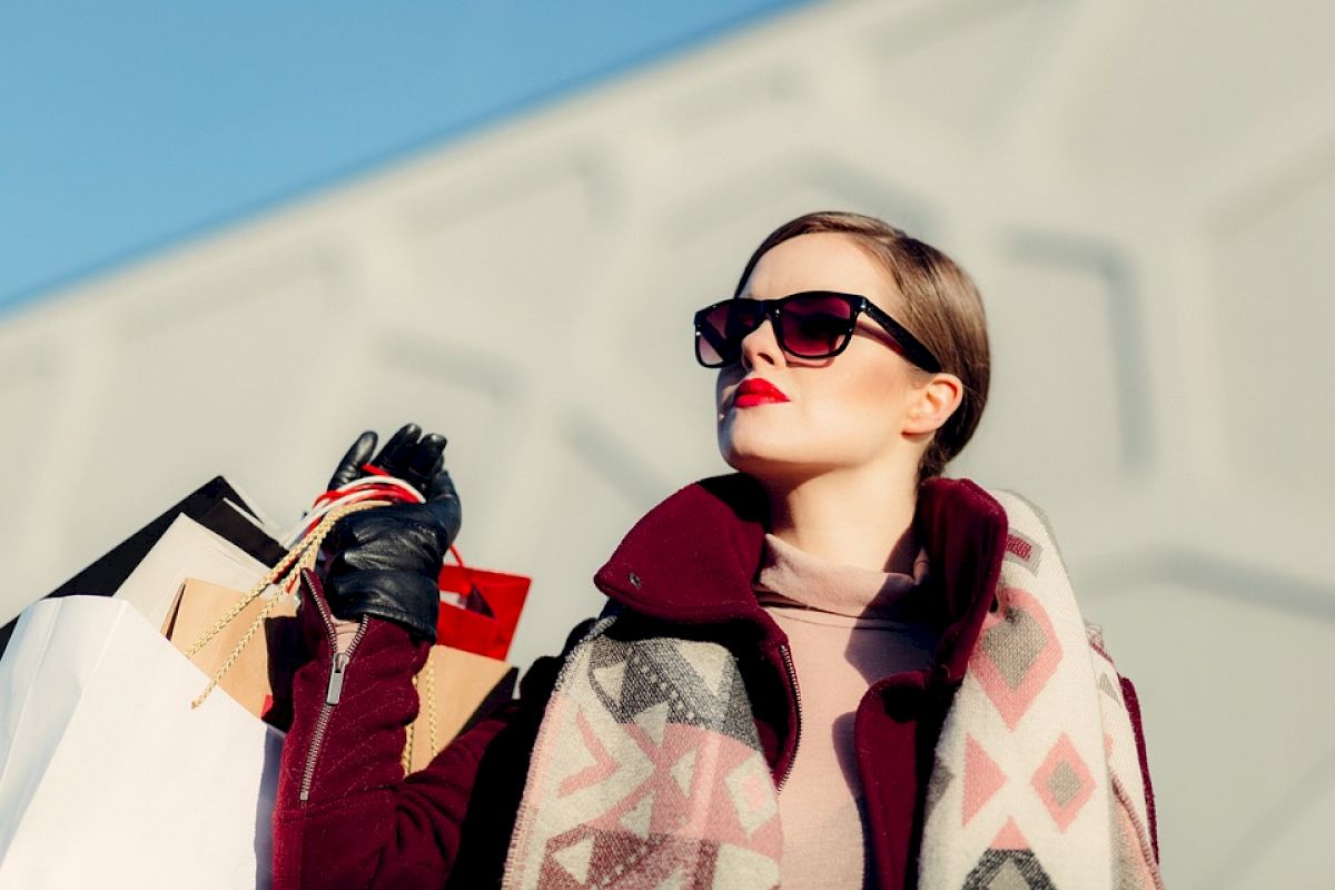 A stylish person wearing sunglasses and holding shopping bags against an urban backdrop. They are dressed in a scarf and a coat.