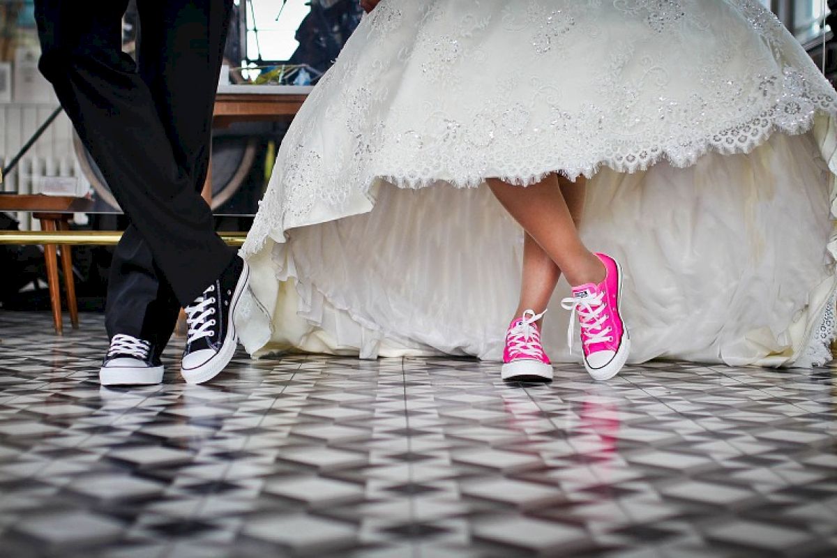 A bride and groom wearing sneakers, with the bride in a white dress and pink shoes, and the groom in black pants and black shoes, stand on a tiled floor.