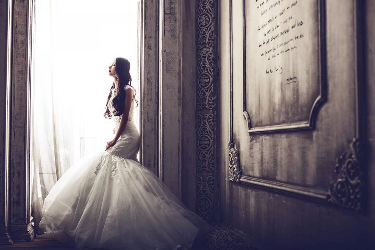 A bride in a white gown stands by a window, gazing outside, with an ornate framed piece of writing on the wall next to her.