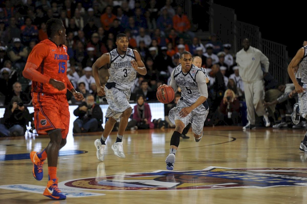 The image shows a college basketball game in action, with players in gray camo and orange uniforms on the court, and a crowd in the background.