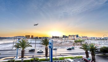 A plane flies over a cityscape with a Ferris wheel, palm trees, and roads in the foreground, under a vivid sunset.