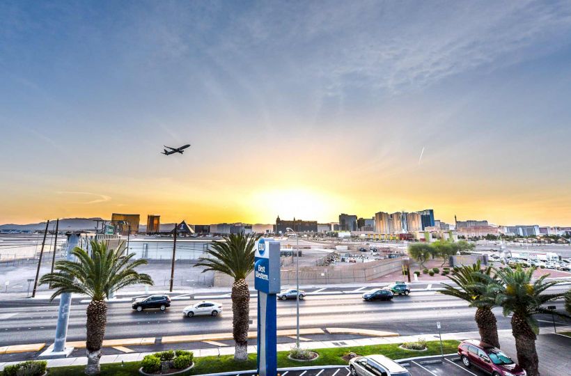 A plane flies over a cityscape with a Ferris wheel, palm trees, and roads in the foreground, under a vivid sunset.