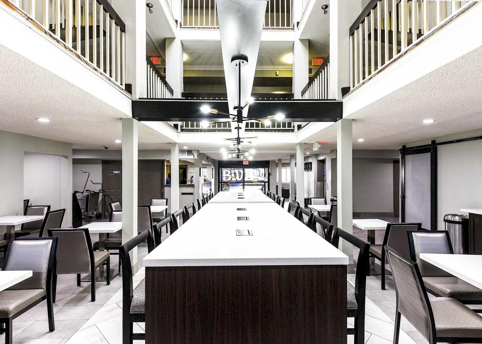The image shows a modern dining area with a long central table, surrounded by chairs, and upper-level balconies with railings in the background.
