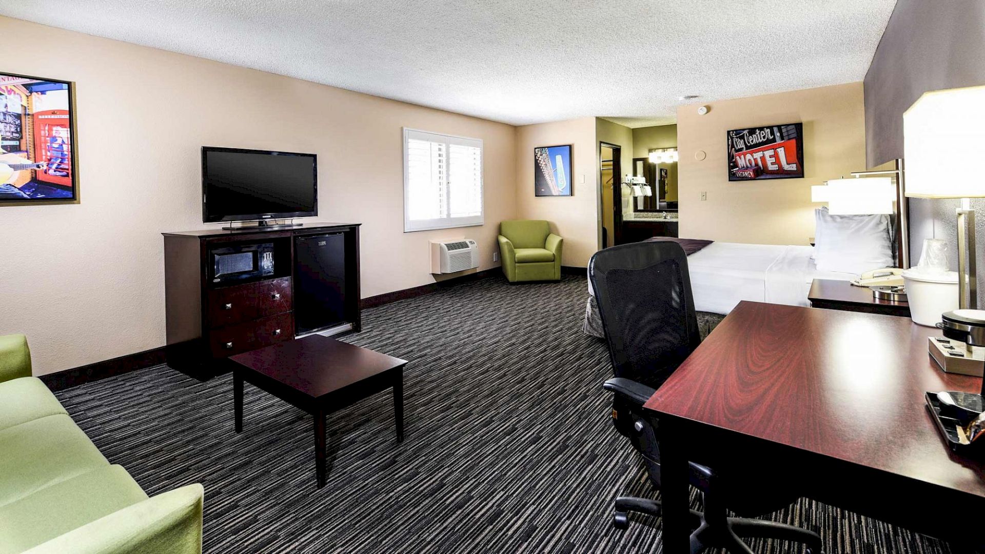 The image shows a hotel room with a bed, desk, chair, TV, sofa, and artwork on the walls, creating a cozy atmosphere.