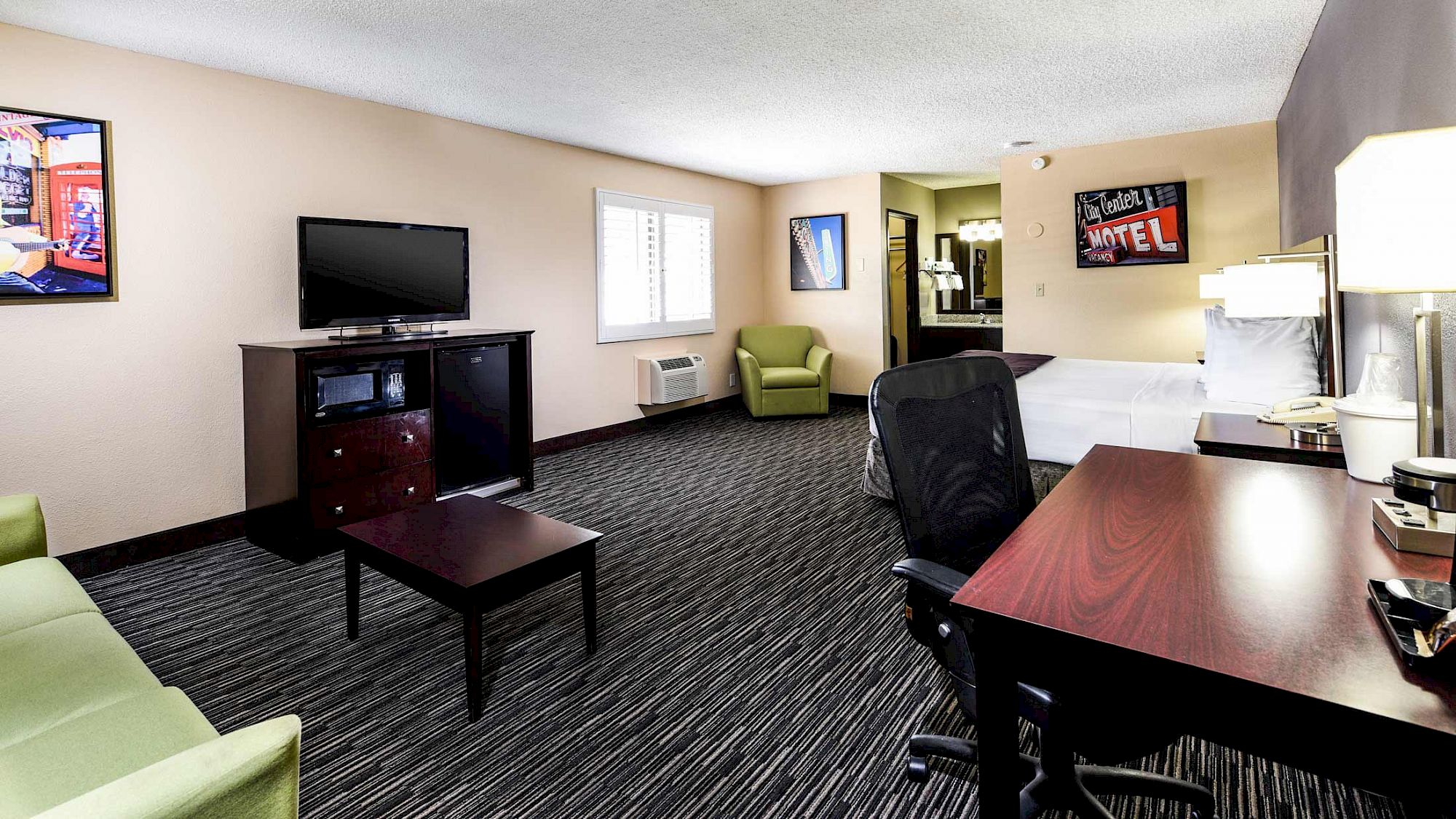 The image shows a hotel room with a bed, desk, chair, TV, sofa, and artwork on the walls, creating a cozy atmosphere.