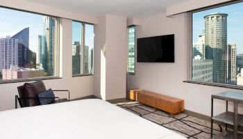 A modern hotel room with large windows showcasing a city skyline, a wall-mounted TV, armchair, and minimalistic furniture, ending the sentence.