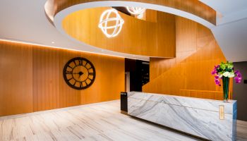 The image shows a modern lobby with a marble reception desk, a large wall clock, decorative lighting, and a floral arrangement on the desk.