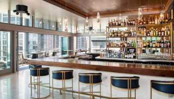 A modern bar with a sleek, wooden counter, blue bar stools, and a well-stocked liquor shelf, featuring large windows with a view of a city.