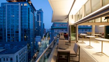 A modern urban balcony at twilight with outdoor seating overlooks a street lined with tall buildings and lit windows ending the sentence.