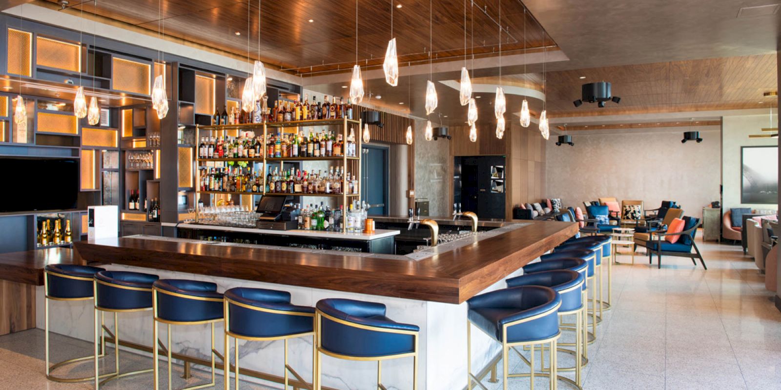 The image depicts a modern bar area with stylish stools, a well-stocked bar, pendant lighting, and a cozy seating area in the background.