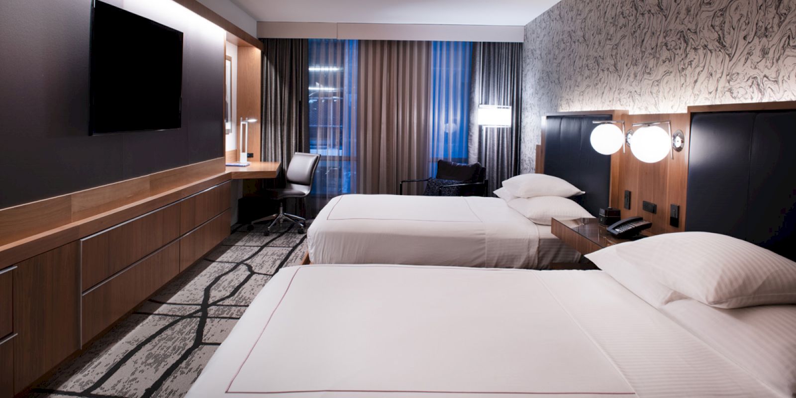 The image shows a modern hotel room with two neatly made beds, a desk, chair, flat-screen TV, and contemporary decor and lighting fixtures.