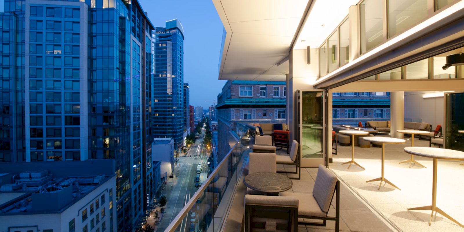 The image shows a cityscape during twilight, with modern high-rise buildings and a spacious, well-lit balcony with tables and chairs.