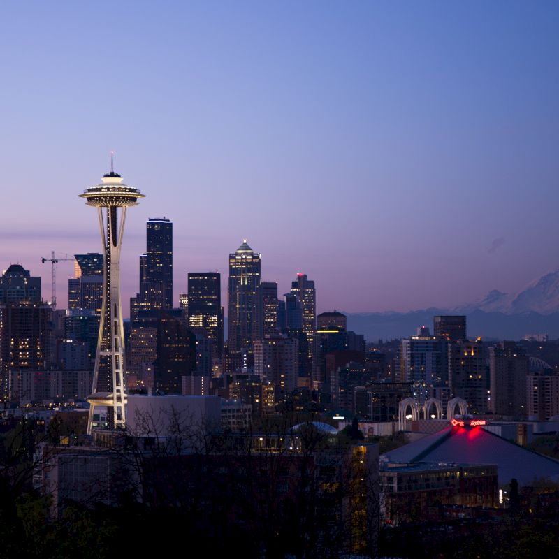Seattle skyline at dusk, featuring the Space Needle with Mount Rainier visible in the background and city lights beginning to shine.