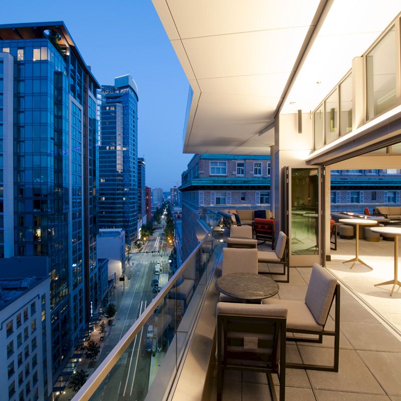 A modern cityscape at twilight with tall buildings and a rooftop terrace featuring tables, chairs, and ambient lighting.