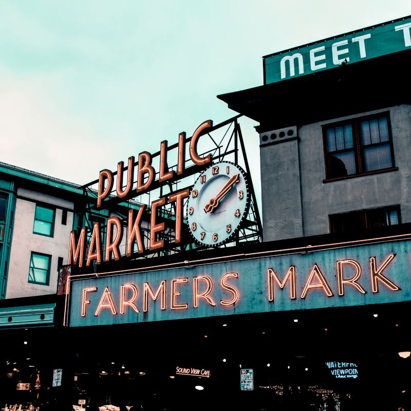 The image shows a public market with neon signs reading "Public Market" and "Farmers Market." An iconic clock is also visible in front of the market.
