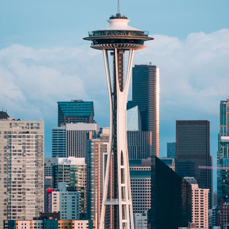 The image shows the iconic Space Needle with the Seattle skyline in the background, featuring various high-rise buildings and a partly cloudy sky.