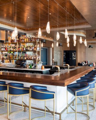 This image shows a modern, well-lit bar with wooden decor, blue bar stools, hanging light fixtures, and a variety of bottles displayed behind the counter.