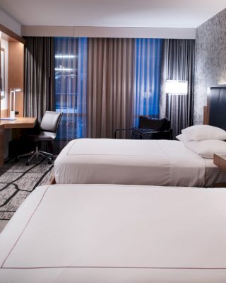A modern hotel room features two neatly made beds, a flat-screen TV, a desk with a chair, and stylish decor, including patterned wallpaper and lighting.