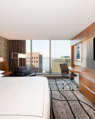 A modern hotel room with a large bed, a desk with a chair, a flat-screen TV, and a window showcasing a cityscape view.