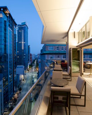 A modern cityscape at dusk with tall buildings and a balcony featuring tables and chairs, overlooking the street below.