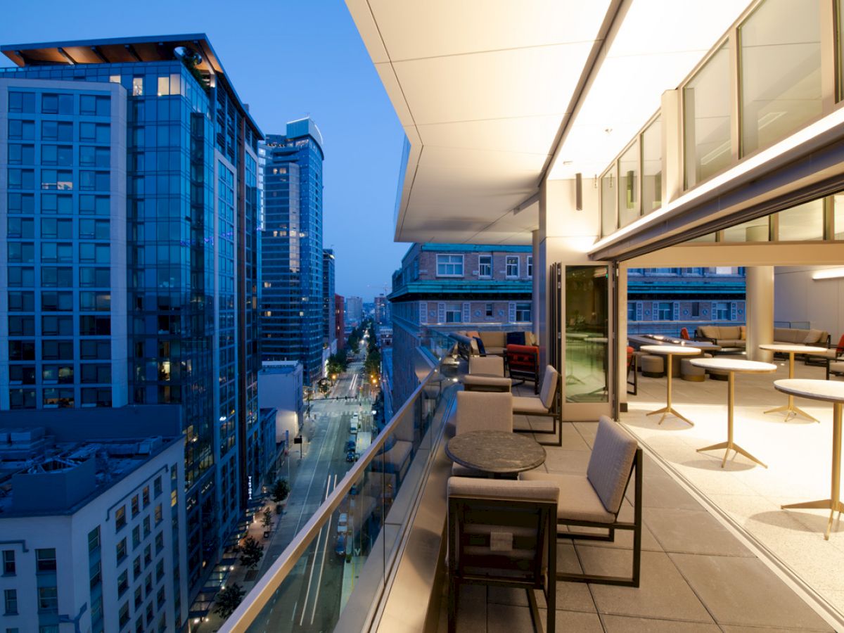 A modern balcony with seating and tables overlooks a city street lined with tall buildings, captured during dusk or early evening.