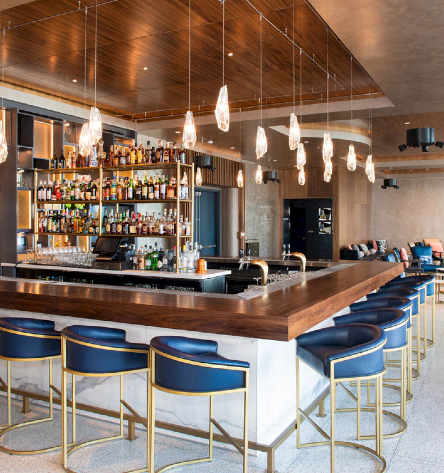 A modern, stylish bar with wooden accents, blue bar stools, and numerous bottles on shelves. Pendant lights hang from the ceiling.
