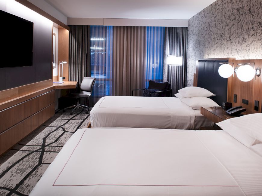 A modern hotel room with two beds, a flat-screen TV, a desk with a chair, and large windows with curtains in a stylishly decorated interior.