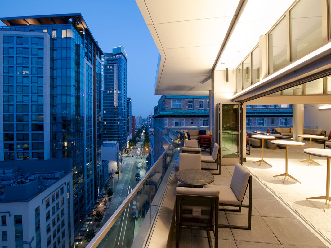 The image shows a modern balcony view at dusk, overlooking a cityscape with tall buildings and a street below. The balcony has seating and tables.