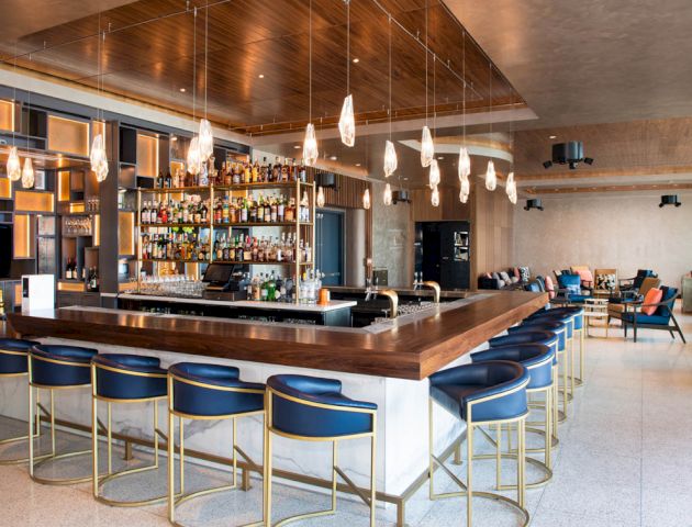 The image shows a modern bar with a wooden counter, blue cushioned bar stools, hanging lights, and a well-stocked alcohol display in a spacious setting.