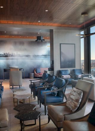 The image shows a modern lounge with comfortable seating, large windows with a scenic view of water and a Ferris wheel.