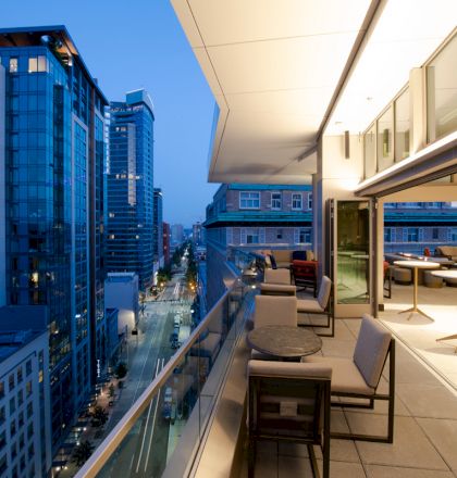 A modern balcony overlooking a cityscape at dusk, with seating and tables under a canopy, and skyscrapers illuminated in the background.