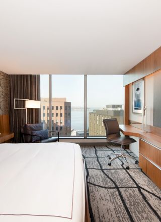 Modern hotel room with a large bed, a wall-mounted TV, a desk with a chair, and a view of city buildings through a large window.