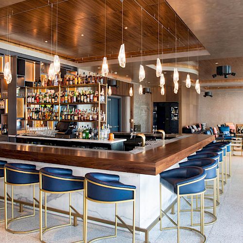 The image shows a modern bar with a wooden counter, blue stools, shelves of alcohol, and hanging pendant lights. Seating and decor are visible.