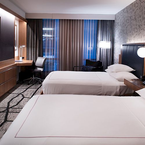 A modern hotel room with two double beds, a wall-mounted TV, a desk with a chair, and stylish lighting and decor.