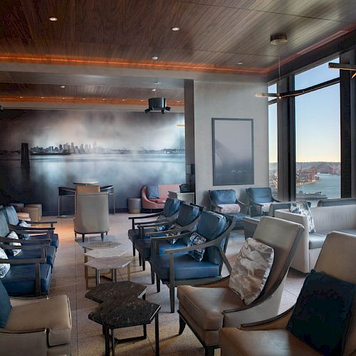 The image depicts a modern, well-lit lounge with armchairs, large windows offering a scenic view of water and boats, and a mural on the back wall.