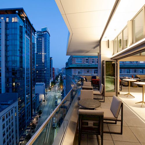 A modern cityscape at dusk, with high-rise buildings and a rooftop terrace featuring seating arrangements and tables overlooking the street.